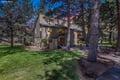 A peaceful mountain retreat nestled among tall pines on a manicured lawn. The building features warm lighting and balconies amid natural surroundings, with a colorful sunset sky as backdrop.